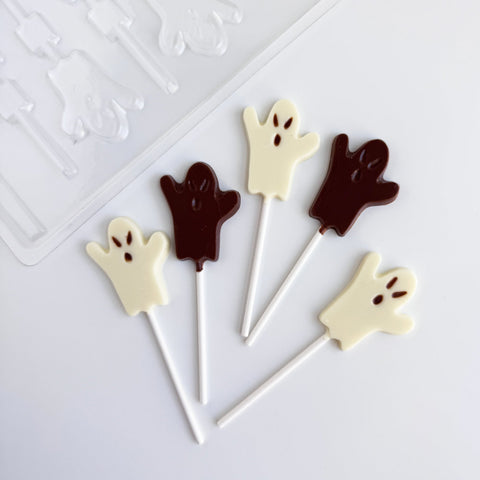 Five ghost-shaped lollipops in white and dark chocolate, arranged on a white surface with a clear plastic candy mold partially visible in the top left corner.