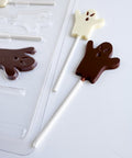 Two ghost-shaped lollipops, one milk chocolate and one white, displayed next to a clear plastic ghost-shaped chocolate mold on a white background.