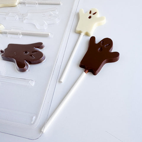 Two ghost-shaped lollipops, one milk chocolate and one white, displayed next to a clear plastic ghost-shaped chocolate mold on a white background.