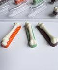 Three bone-shaped chocolate pieces in orange, green, and dark brown with white bone shaped centers, arranged on a white surface with more chocolate molds in the background.