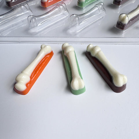 Three bone-shaped chocolate pieces in orange, green, and dark brown with white bone shaped centers, arranged on a white surface with more chocolate molds in the background.