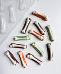 Small bone-shaped chocolate pieces in white over top of a colorful rectangle base of dark brown, orange, and green scattered on a white surface, with a clear plastic mold tray partially visible in the upper left corner.