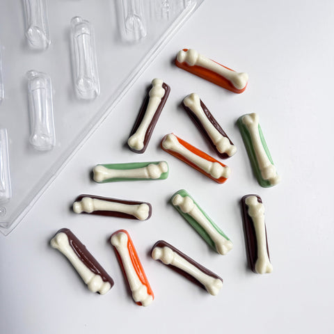 Small bone-shaped chocolate pieces in white over top of a colorful rectangle base of dark brown, orange, and green scattered on a white surface, with a clear plastic mold tray partially visible in the upper left corner.