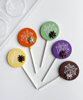 Five Halloween-themed round lollipops with spiderweb and spiders molded into the centers, displayed on a white background with the plastic  candy mold used to make them partially visible in the top left. The lollipops are molded with yellow, orange, green, purple, and milk chocolate.