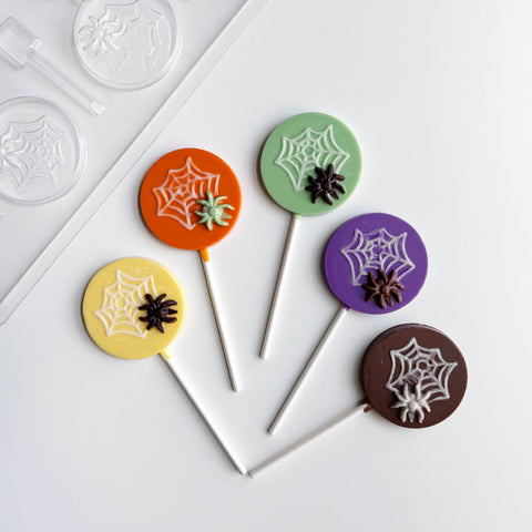 Five Halloween-themed round lollipops with spiderweb and spiders molded into the centers, displayed on a white background with the plastic  candy mold used to make them partially visible in the top left. The lollipops are molded with yellow, orange, green, purple, and milk chocolate.