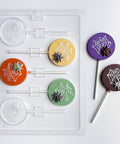 Plastic candy mold that makes round lollipop with spider and web molded into the center. Two finished lollipops to the right. One molded with purple chocolate for the circle, milk chocolate for the spider, and white chocolate for the web. The other molded with dark chocolate for the circle, white chocolate for the web and spider.