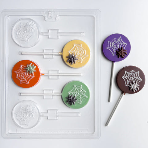 Plastic candy mold that makes round lollipop with spider and web molded into the center. Two finished lollipops to the right. One molded with purple chocolate for the circle, milk chocolate for the spider, and white chocolate for the web. The other molded with dark chocolate for the circle, white chocolate for the web and spider.