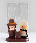 thanksgiving centerpiece chocolate mold with the finished chocolate centerpiece in front of it. The centerpiece features two pilgrims, one female and one male and a turkey in front of them sitting on a base. The clear plastic mold is placed behind it.