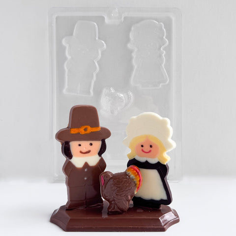 thanksgiving centerpiece chocolate mold with the finished chocolate centerpiece in front of it. The centerpiece features two pilgrims, one female and one male and a turkey in front of them sitting on a base. The clear plastic mold is placed behind it.