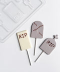 Three tombstone-shaped lollipops with 'RIP' text in dark brown, displayed on a white surface next to a clear plastic Halloween chocolate mold.