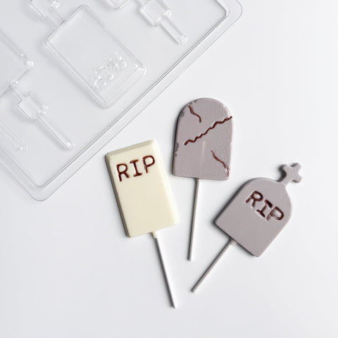 Three tombstone-shaped lollipops with 'RIP' text in dark brown, displayed on a white surface next to a clear plastic Halloween chocolate mold.