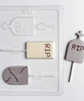 A Halloween-themed chocolate mold on a white background. Featuring tombstone shaped lollipops in three different designs., one rectangle with straight edges and RIP molded into the center, one rounded classic grave stone shape with a cracked look, and one rounded with a cross shape protruding off the top and RIP molded into the center. Shown with a finished lollipop to the right.