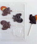 A clear plastic turkey-shaped lollipop mold shown on a white background, alongside a finished lollipop made with dark chocolate with orange, red, and yellow chocolate to accent the tail.