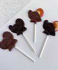 Four turkey-shaped lollipops on white sticks, made with dark chocolate and milk chocolate, arranged on a white surface with a clear plastic candy mold partially visible in the top left.