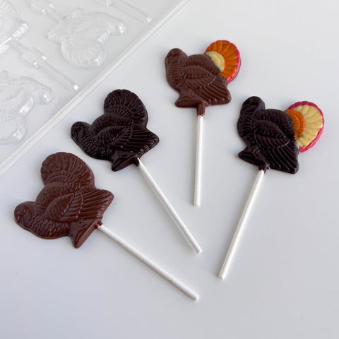 Four turkey-shaped lollipops on white sticks, made with dark chocolate and milk chocolate, arranged on a white surface with a clear plastic candy mold partially visible in the top left.