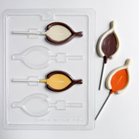 A clear plastic thanksgiving lollipop candy mold shaped like a wishbone. Shown along side finished chocolate wishbone lollipops made from white chocolate, milk chocolate, yellow, and orange colored chocolate.