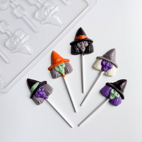Five Halloween-themed witch head chocolate lollipops with witch hats and faces, arranged on a white surface next to a clear plastic candy mold.