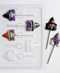 Witch face chocolate lollipops with three inside of the plastic candy mold used to make them and two sitting outside of the mold to the right. The lollipops are made with an assortment of colored chocolate.